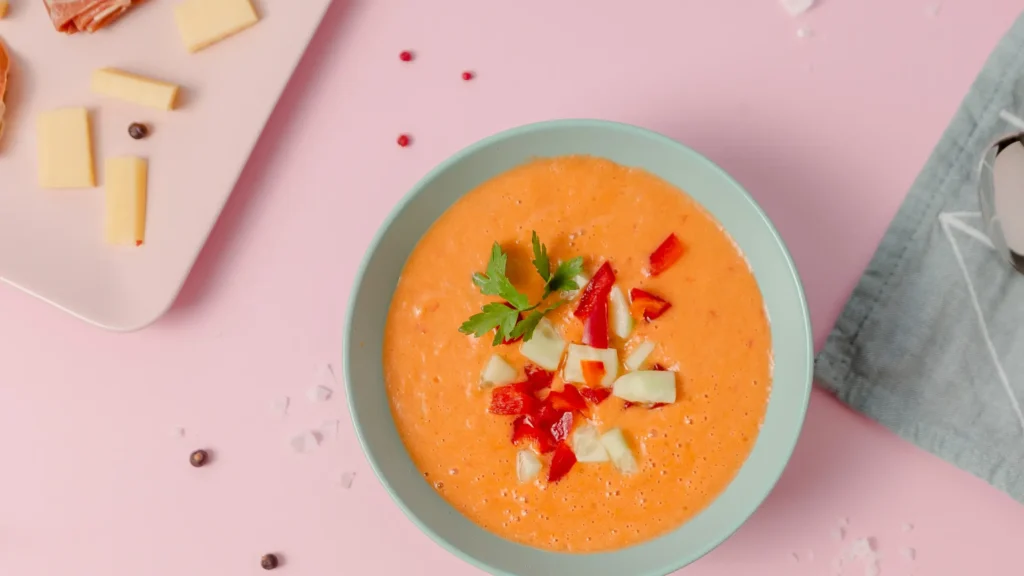 bowl of gazpacho on a pink table with cheese and ham