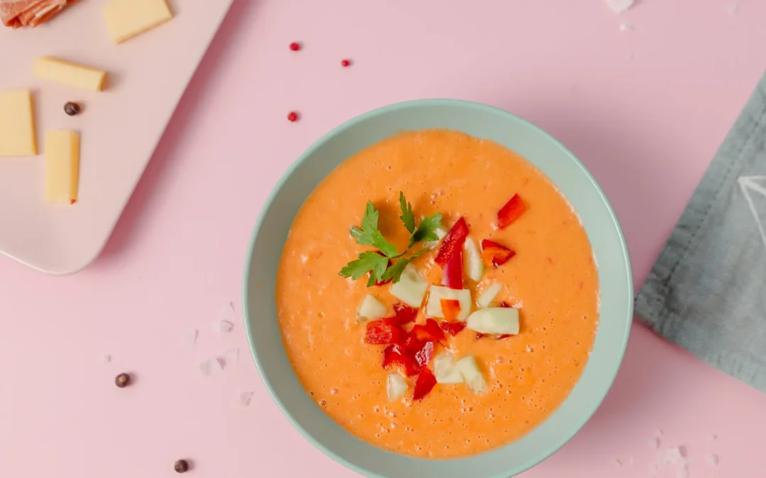 bowl of gazpacho on a pink table with cheese and ham