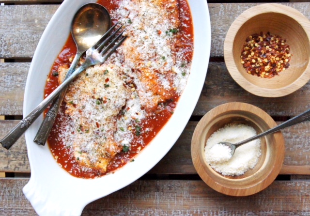 chicken parmesan in a white dish with small wood bowls filled with parmesan cheese and red chili peppers