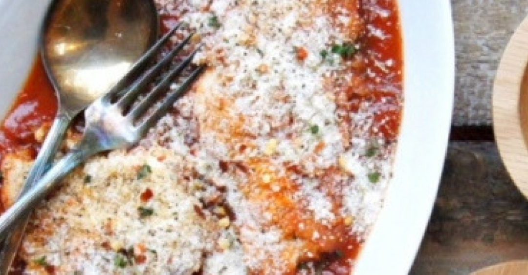 chicken parmesan in a white serving dish with a fork and a side of red chili peppers