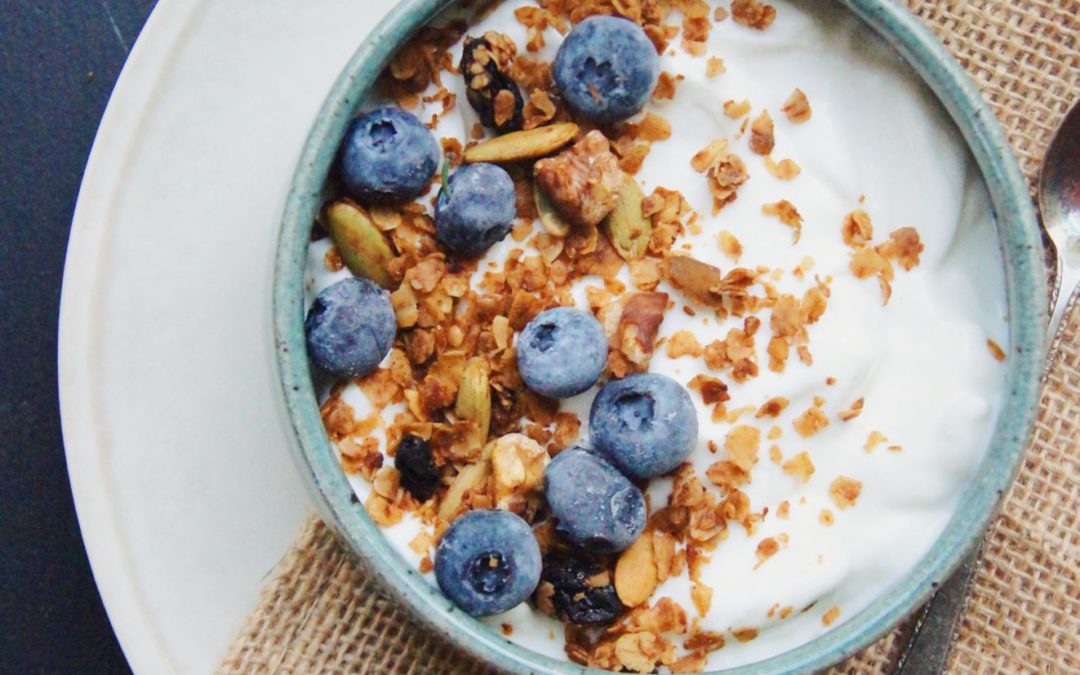 A bowl of yogurt with granola and blueberries. A spoon is to the right of the bowl.