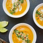 three bowls of carrot and coconut curry soup garnished with herbs and red chili peppers, limes are on the side