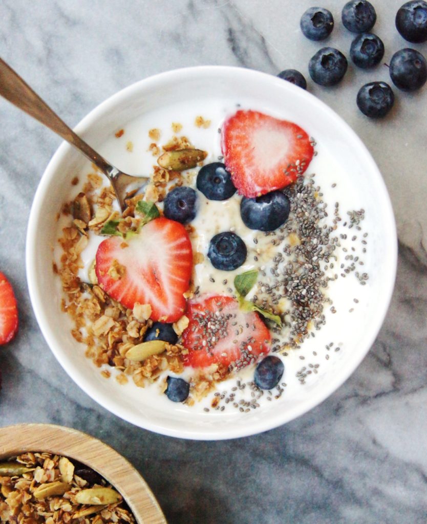 Bowl of plain yogurt with chia seeds, granola, strawberries and blueberries with a spoon in yogurt, berries on the side of the bowl