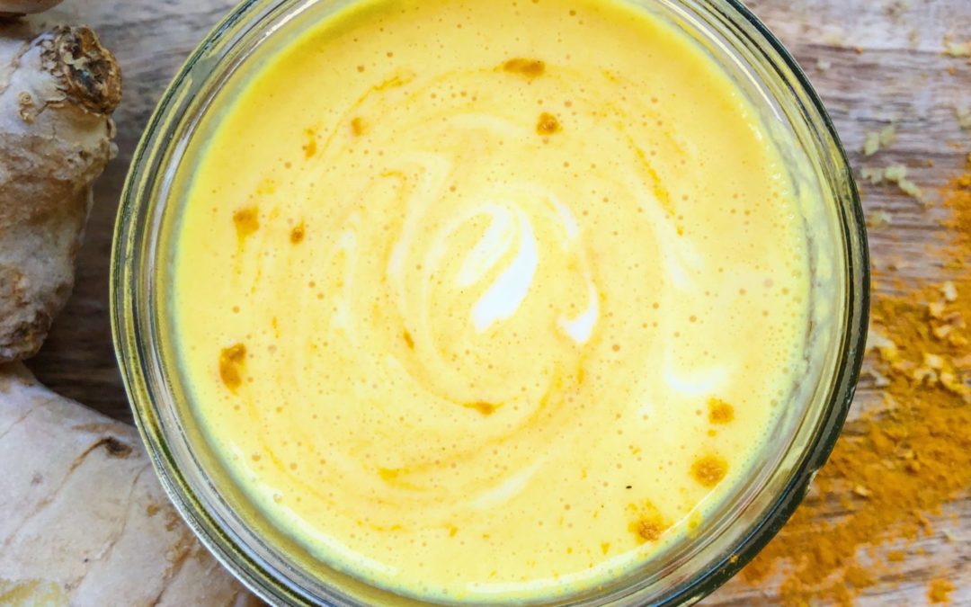 overhead shot of turmeric and ginger smoothie in a clear glass with turmeric powder on wood plank and ginger root to left of smoothie