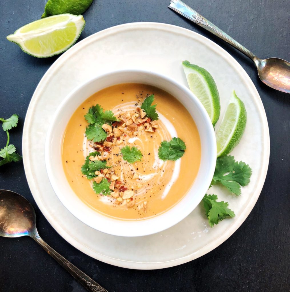 Bowl of squash soup garnished with ground peanuts, limes, cilantro and red chili peppers, two spoons beside the bowls