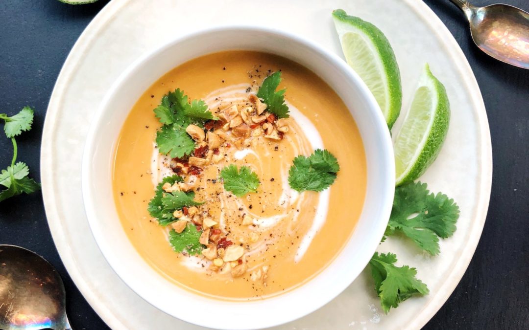Bowl of squash soup garnished with ground peanuts, limes, cilantro and red chili peppers, two spoons beside the bowls