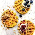 3 waffles with topped with blueberries, pomegranate seeds, nut butter and greek yogurt + hemp and flax seeds on a white cutting board