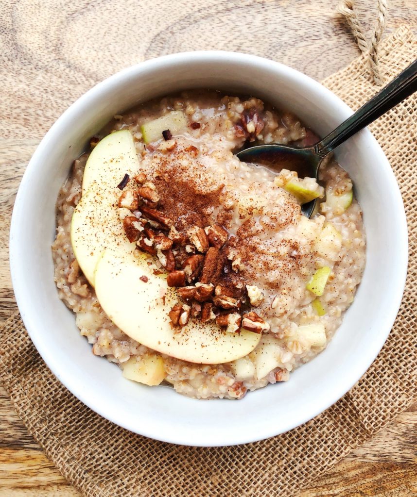 bowl of apple oatmeal shown in a white bowl with cinnamon and pecans as a topping, a spoon is coming out on the right side of bowl