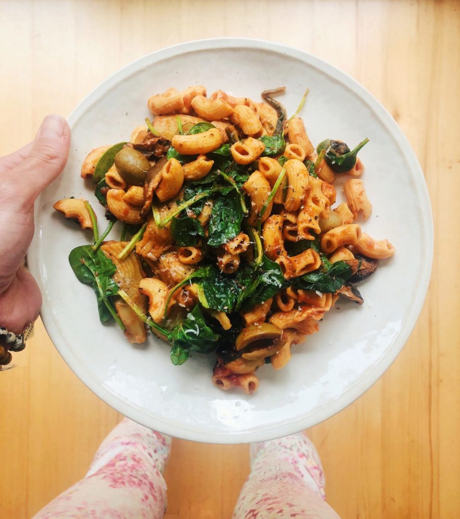 plate of banza chickpea pasta on a white plate with spinach, olives and hand of girl holding the plate