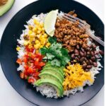 black bowl with black beans, avocado, rice, peppers, corn, cheese, taco meat, and a lime